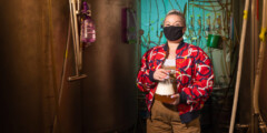 Ellie Hadley, wearing a bright red patterned jacket, stands in a brewery in front of pipes and fermenters. She smiles under her mask.