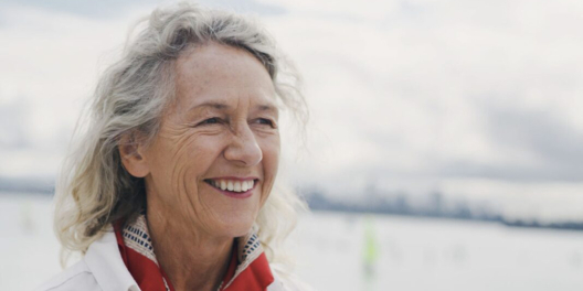 Joyce Murray smiling on a cloudy day. Her hair is grey and she is wearing a small red scarf around her neck.