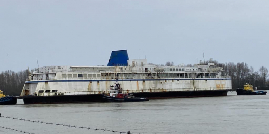 An old ferry sits rusting on the shore against a bleak backdrop.