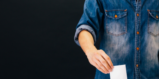 A closeup of someone placing a ballot in a box. The person is wearing a blue denim shirt. The photo is cropped to show only the torso and the hand with the ballot.