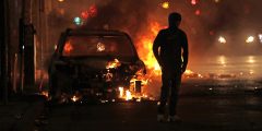 A person walks toward the camera and away from a burning car. It's dark and their face is covered.